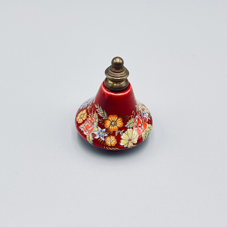May include: A bell-shaped ceramic object with a glossy red finish, decorated with a colorful floral pattern featuring orange, yellow, and blue flowers. The top is capped with a metallic, tiered finial in a muted brass tone.