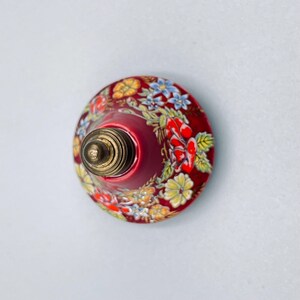 May include: A round, dome-shaped knob featuring a deep red base color decorated with a vibrant floral pattern of yellow, blue, and red flowers with green leaves. The center of the knob has a metallic, brass-toned threaded screw attachment.