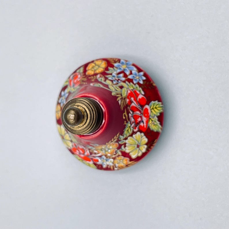 May include: A round, dome-shaped knob featuring a deep red base color decorated with a vibrant floral pattern of yellow, blue, and red flowers with green leaves. The center of the knob has a metallic, brass-toned threaded screw attachment.