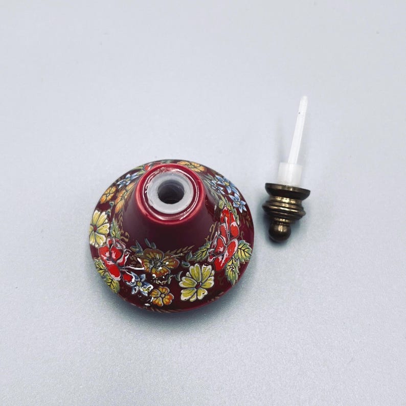 May include: A conical ceramic bottle with a deep red base color, decorated with a hand-painted floral pattern featuring yellow, red, and green blossoms. The bottle has a central opening and comes with a separate metallic stopper with a white stem.