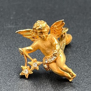 May include: A gold-toned cherub brooch with outstretched wings and holding two star charms. The cherub's sash is adorned with small, clear gemstones. The brooch is set against a dark background.