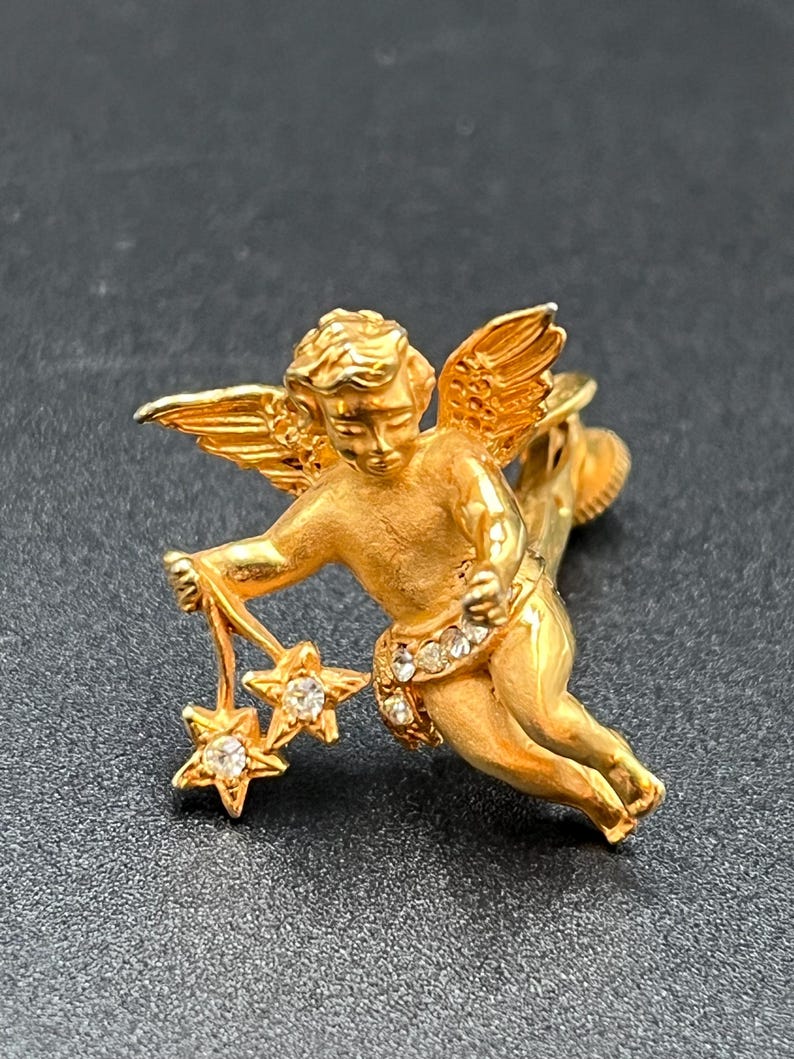 May include: A gold-toned cherub brooch with outstretched wings and holding two star charms. The cherub's sash is adorned with small, clear gemstones. The brooch is set against a dark background.