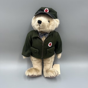 May include: A plush teddy bear with cream-colored fur wearing a dark green zip-up jacket with two front pockets and a red star logo. The bear wears a matching dark green cap with a red star emblem and a small blue patterned bowtie at the neck.