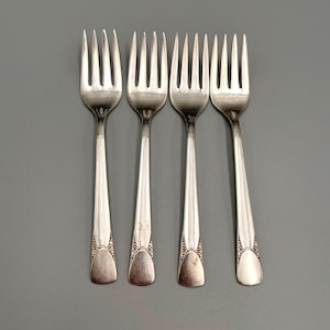 Pode incluir: Quatro garfos prateados com dentes e cabos decorativos. Os garfos estão dispostos em fila, com os dentes voltados para cima. Os cabos têm um design detalhado perto da base. Os garfos são feitos de metal brilhante, sobre fundo cinza.