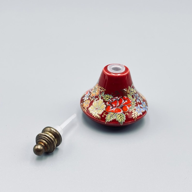 May include: A red ceramic bottle stopper with a floral pattern featuring red roses and green leaves with gold accents. The stopper has a conical shape and includes a separate metallic-colored base piece with a white plastic stem for insertion.