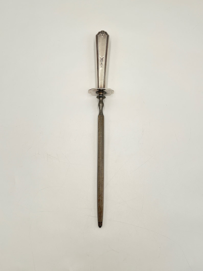 May include: A silver knife sharpener with a decorative handle and a round base. The sharpener is made of metal and has a long, thin blade.