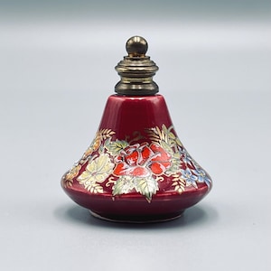 May include: A small, conical, deep red ceramic perfume bottle featuring a hand-painted floral design with red roses, green leaves, and blue accents. The bottle is topped with a metallic, tiered, spherical-topped stopper in a muted gold tone.