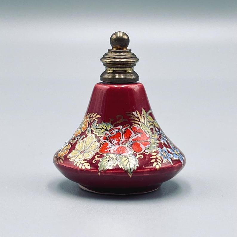 May include: A small, conical, deep red ceramic perfume bottle featuring a hand-painted floral design with red roses, green leaves, and blue accents. The bottle is topped with a metallic, tiered, spherical-topped stopper in a muted gold tone.