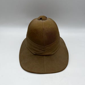 May include: A brown pith helmet with a wide brim and a decorative band around the crown. The hat has a rounded top and a small, rounded peak. The hat is made of a light-colored material.