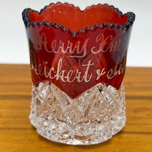 May include: A red glass cup with a scalloped rim and a clear, cut-glass base. The cup has the inscription "Henry Emma Reichert & Co." in white lettering.