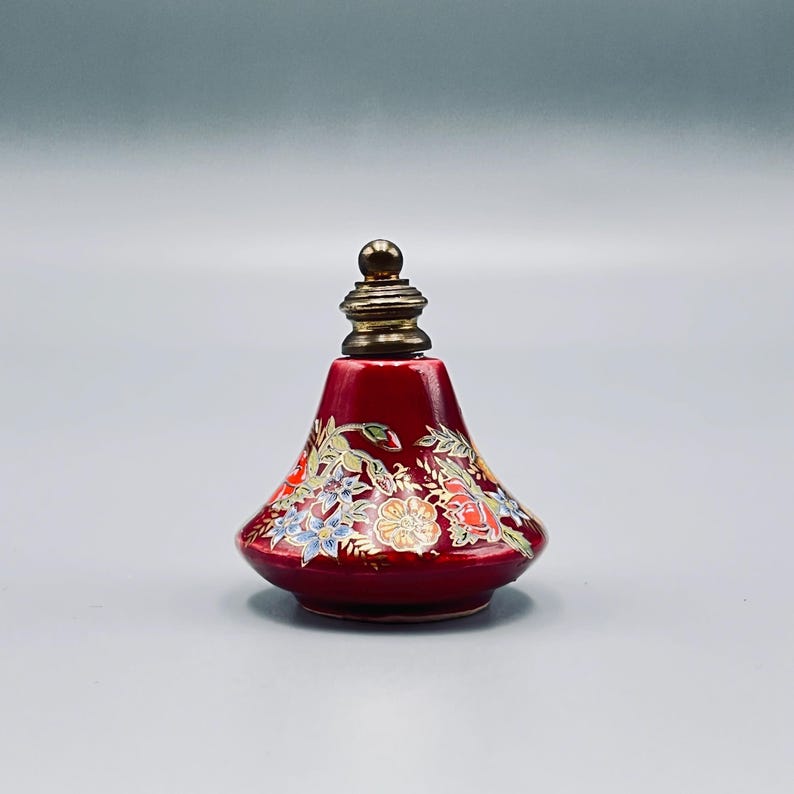 May include: A small, conical red perfume bottle featuring a metallic gold-toned stopper. The body of the bottle is decorated with a colorful floral pattern in shades of blue, orange, and green, accented with fine gold-colored line details.