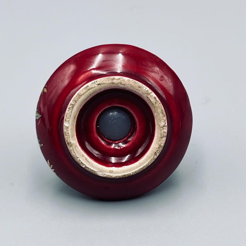 May include: A circular, deep red ceramic knob with a glossy finish. The underside features a central circular indentation with a dark gray plug and a beige, unglazed rim. The side of the knob displays a small, colorful floral pattern.