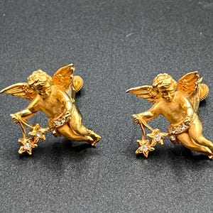May include: A pair of gold-colored angel earrings. Each earring features a cherub holding a star-shaped charm with clear crystals. The angels have detailed wings and are posed in a dynamic, flowing position. The earrings are set against a dark background.