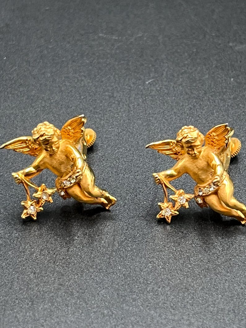 May include: A pair of gold-colored angel earrings. Each earring features a cherub holding a star-shaped charm with clear crystals. The angels have detailed wings and are posed in a dynamic, flowing position. The earrings are set against a dark background.