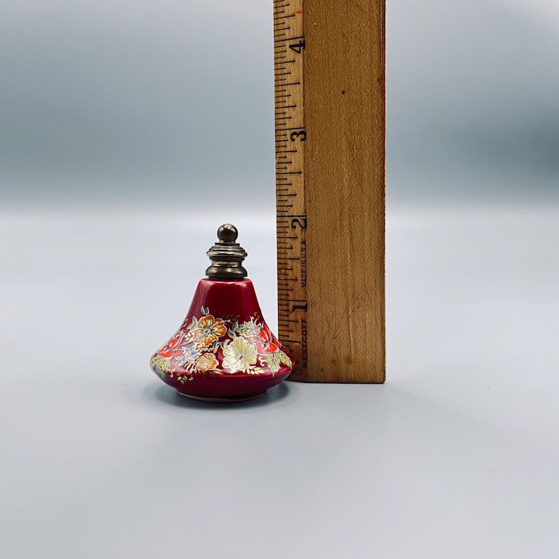 May include: A small, bell-shaped red ceramic perfume bottle with a metallic stopper, decorated with a multi-colored floral pattern. A wooden ruler placed next to the bottle indicates a height of approximately 1.75 inches for the bottle.