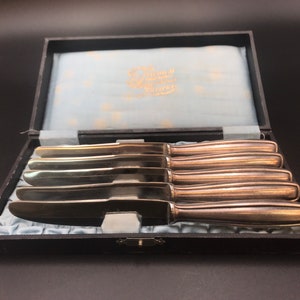 May include: A set of six silver-colored knives in a black box with a light blue satin lining. The knives have a classic design with a rounded handle and a sharp blade. The box has gold lettering on the inside.