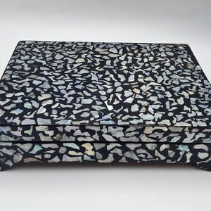 May include: A rectangular black jewelry box with a mosaic pattern of white and iridescent shell pieces. The box has a hinged lid and rests on four small feet, offering a decorative storage solution.