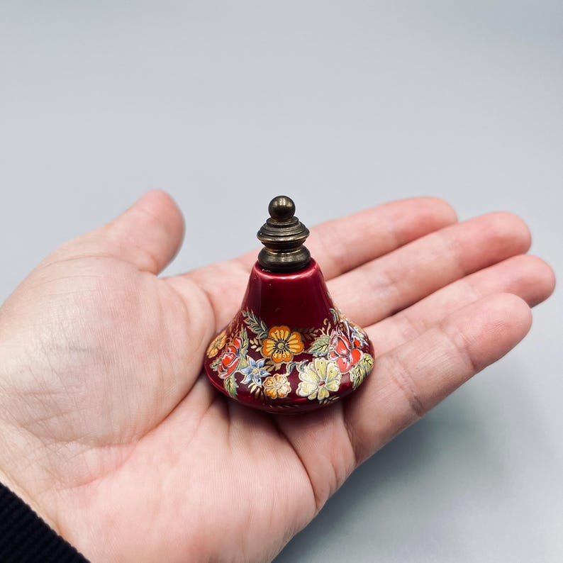 May include: A small, bell-shaped, deep red ceramic bottle with a metallic brass-colored stopper. The surface is decorated with a colorful, intricate floral pattern featuring orange, yellow, and white blossoms with green leaves around the base.