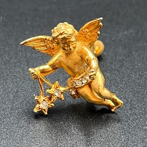 May include: A gold-colored cherub pin with outstretched wings and a flowing pose. The cherub holds a string of three star-shaped charms, each with a central clear stone. The cherub's waist is adorned with a band of clear stones.