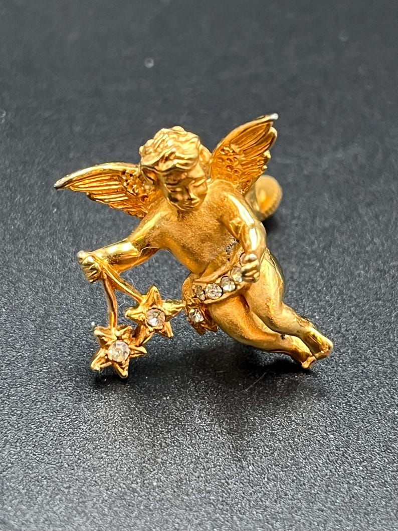 May include: A gold-colored cherub pin with outstretched wings and a flowing pose. The cherub holds a string of three star-shaped charms, each with a central clear stone. The cherub's waist is adorned with a band of clear stones.
