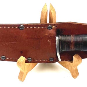 Puede incluir: Una funda de cuero marrón con borde remachado contiene un cuchillo con mango negro y hoja plateada. La funda se asegura con una correa de cuero y un botón negro. El cuchillo se exhibe sobre un soporte de madera.