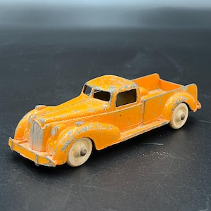 Pode incluir: Um brinquedo antigo de metal fundido em forma de caminhonete pickup laranja, com desgaste visível e lascas na pintura. O veículo apresenta uma grade frontal proeminente, uma cabine com janelas abertas, uma caçamba e quatro rodas brancas.