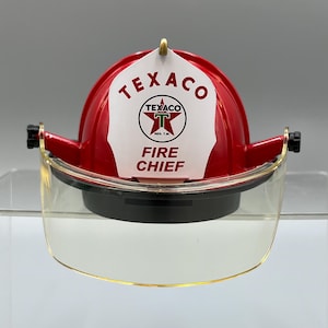 Może przedstawiać: Czerwony hełm strażacki z białą osłoną czołową z logo Texaco i napisem Texaco Fire Chief. Hełm posiada przezroczystą, zakrzywioną plastikową przyłbicę przymocowaną po bokach za pomocą czarnych elementów montażowych i czarną podstawę.