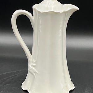 May include: A tall, white ceramic pitcher with a lid. The pitcher has a curved handle and a spout for pouring. The design includes a floral motif on the side and a scalloped edge at the base and lid. The pitcher is approximately 10 inches tall.