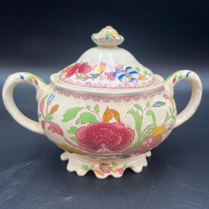 May include: Cream-colored ceramic sugar bowl with a lid, featuring a floral design in red, pink, blue, and green. The bowl has two handles and a scalloped base. The lid is topped with a small, decorative knob.