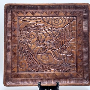 May include: Square wooden tray with a carved dragon design. The tray is brown with a detailed relief carving of a dragon in the center, surrounded by a geometric border. The dragon is depicted with intricate scales and flowing features.