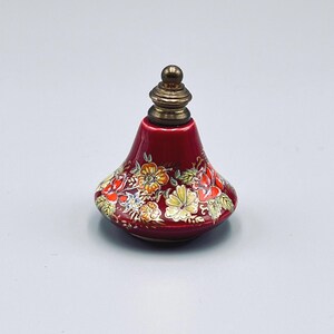 May include: A small, bell-shaped red ceramic perfume bottle featuring a hand-painted floral pattern with yellow, orange, and white blossoms and green leaves. The bottle is topped with a metallic, tiered, brass-colored stopper with a rounded finial.