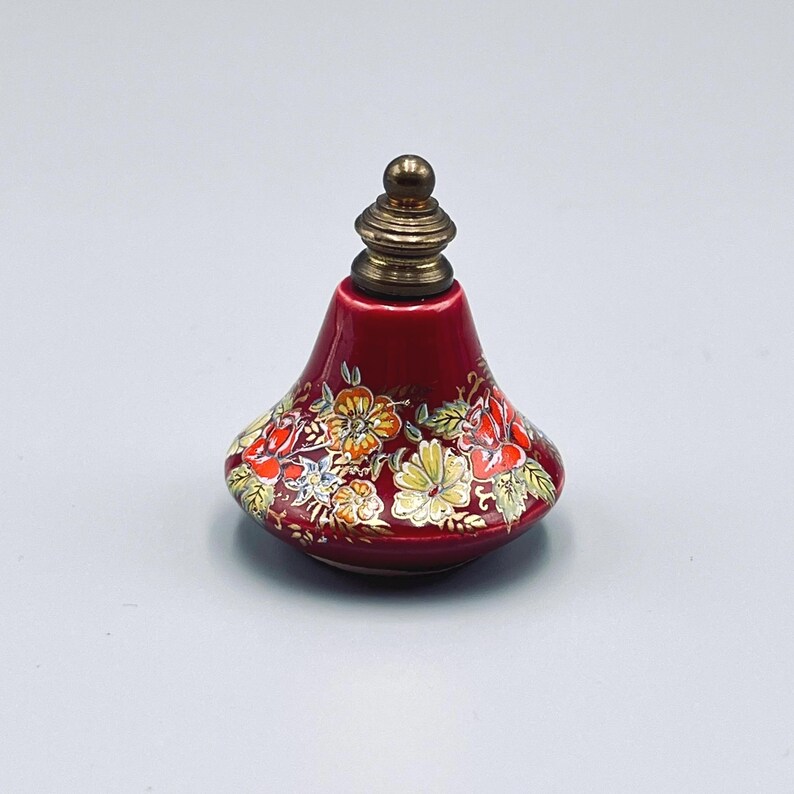 May include: A small, bell-shaped red ceramic perfume bottle featuring a hand-painted floral pattern with yellow, orange, and white blossoms and green leaves. The bottle is topped with a metallic, tiered, brass-colored stopper with a rounded finial.
