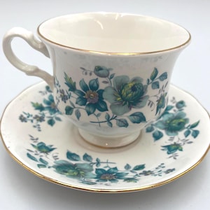 May include: A white teacup and saucer set with a floral design in shades of teal and blue. The cup and saucer have gold trim. The teacup has a curved handle.