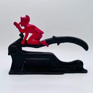 May include: A black and red vintage desk seal with a devil figure on top. The seal has a curved handle and a base with the word "ORIGINAL" embossed on it. The devil figure is red and appears to be holding a small object to its mouth.