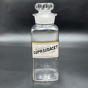 May include: A clear glass bottle with a glass stopper and a label that reads "CUPR.SUBACET."