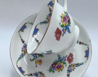 May include: A white porcelain teacup and saucer with a floral pattern in shades of blue, yellow, red, and pink. The cup and saucer have a gold trim around the edges.