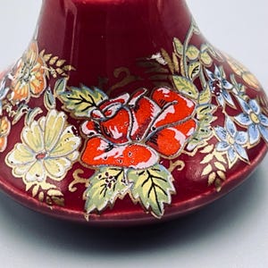 May include: A deep red ceramic bell featuring a hand-painted floral design. The surface is decorated with vibrant orange roses, pale yellow daisies, and light blue blossoms, all accented with delicate gold-colored leaf and vine detailing.