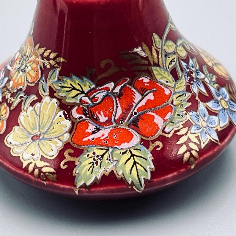 May include: A deep red ceramic bell featuring a hand-painted floral design. The surface is decorated with vibrant orange roses, pale yellow daisies, and light blue blossoms, all accented with delicate gold-colored leaf and vine detailing.
