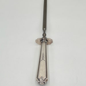 May include: A silver-plated knife sharpener with a long, thin blade and a decorative handle. The handle has a floral design and a small, round knob at the end.