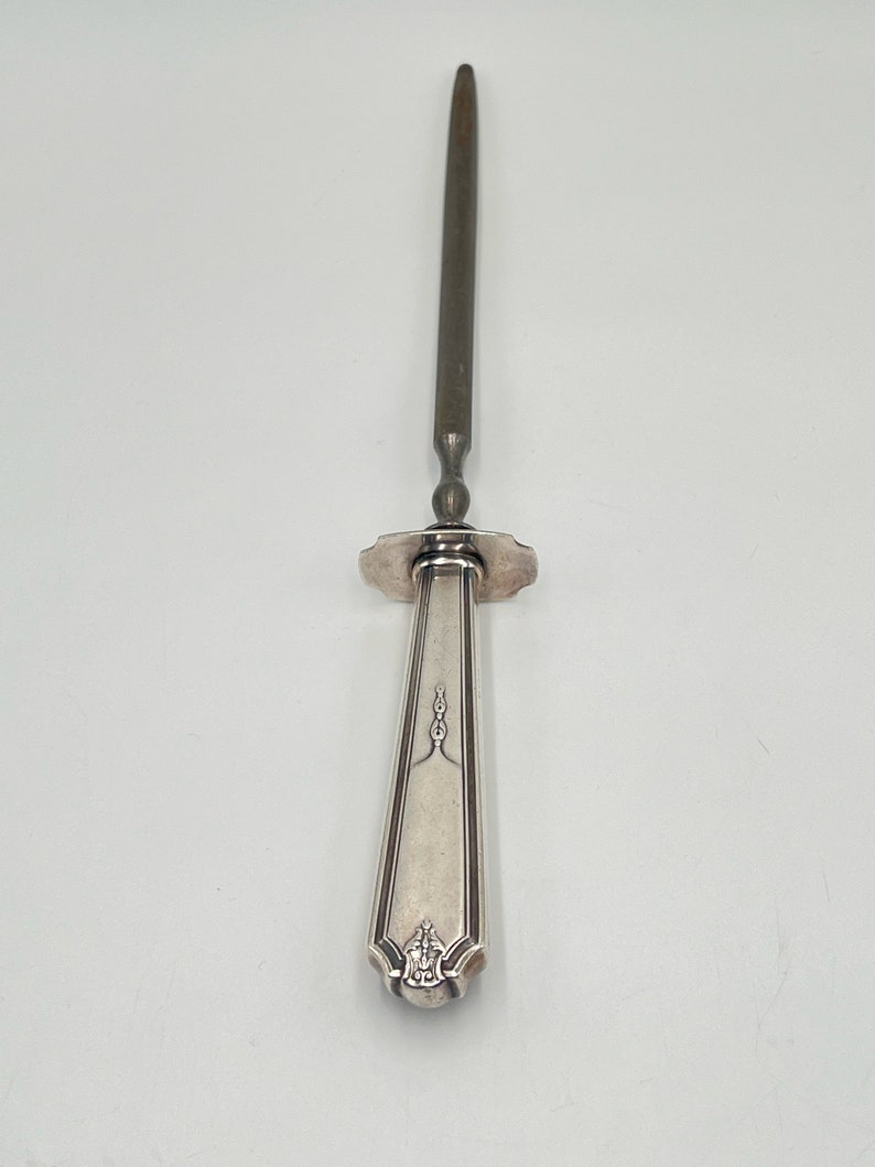 May include: A silver-plated knife sharpener with a long, thin blade and a decorative handle. The handle has a floral design and a small, round knob at the end.