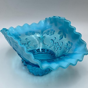May include: A light blue, vintage glass bowl with a ruffled edge and floral design. The bowl has a pedestal base and is made of a translucent glass. The floral pattern is etched into the glass.