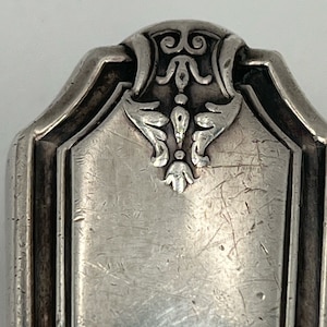 May include: Close-up of the top of a silver flatware piece with an ornate design. The design features a stylized floral motif with a central point and curved lines.