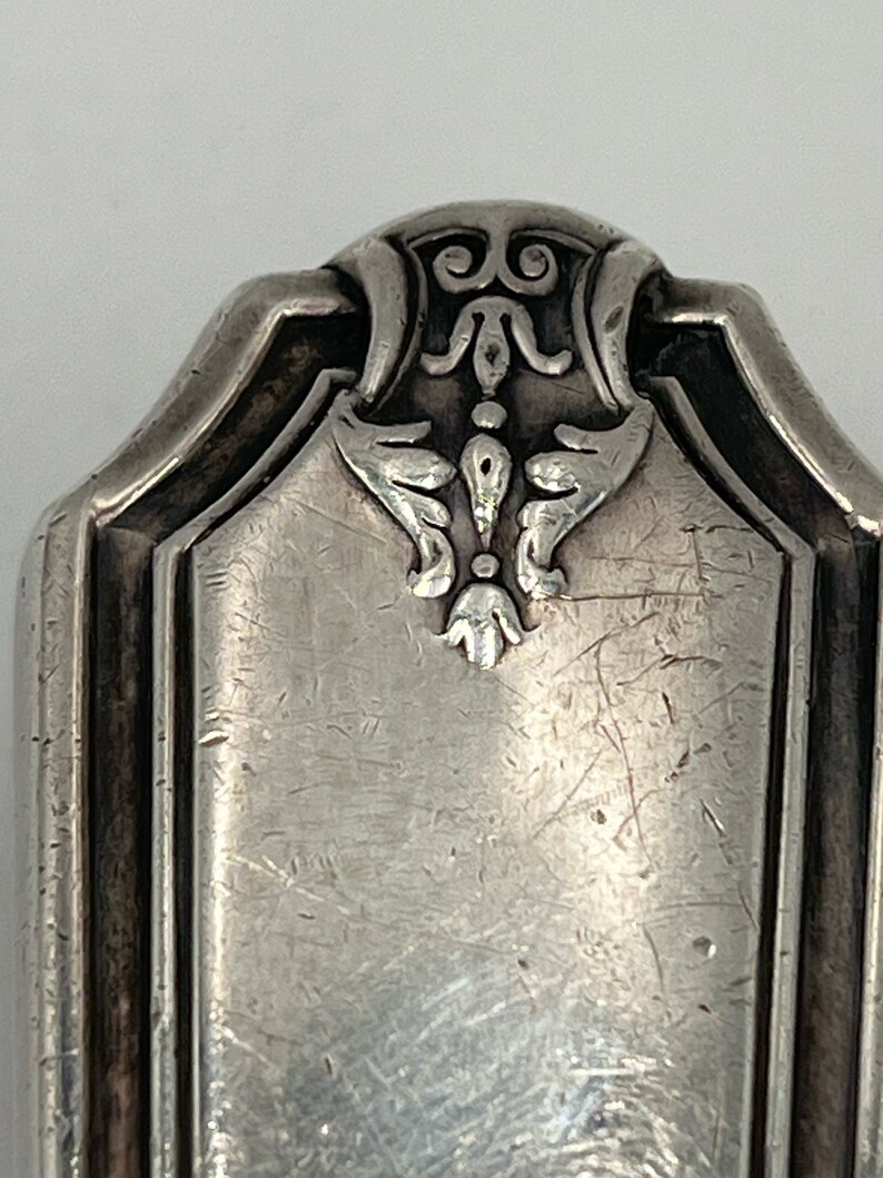 May include: Close-up of the top of a silver flatware piece with an ornate design. The design features a stylized floral motif with a central point and curved lines.