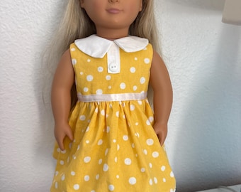 May include: A doll with long blonde hair wearing a sleeveless yellow dress featuring a white polka dot pattern, a white Peter Pan collar, a single white button at the neckline, a thin white ribbon at the waist, and a white lace trim at the hem.
