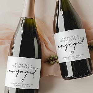 May include: Two dark glass champagne bottles with white labels featuring the black text "PAIRS WELL WITH GETTING engaged" and "CONGRATULATIONS CHLOE & ADAM" with a small heart icon. The bottles have gold foil tops and are positioned side by side.