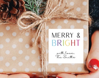 May include: A brown gift box with white polka dots is tied with twine and a sprig of pine. A white gift tag with the text "Merry & Bright" in rainbow colours and "with love, The Smiths" in black text is attached to the box.