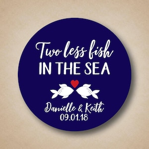 Può includere: Un adesivo rotondo blu navy con testo bianco che recita "Two less fish in the sea" con due sagome di pesci bianchi che si baciano con un cuore rosso in mezzo. Il testo "Danielle & Keith 09.01.18" è sotto i pesci.