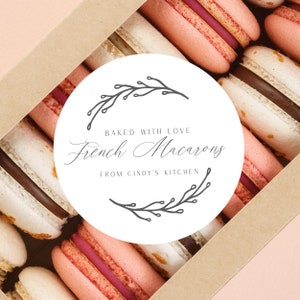 May include: A box of French macarons in various colors, including pink, white, and brown. The macarons are arranged in rows inside a cardboard box. A white circular label with the text "Baked with Love French Macarons From Cindy's Kitchen" is in the center.