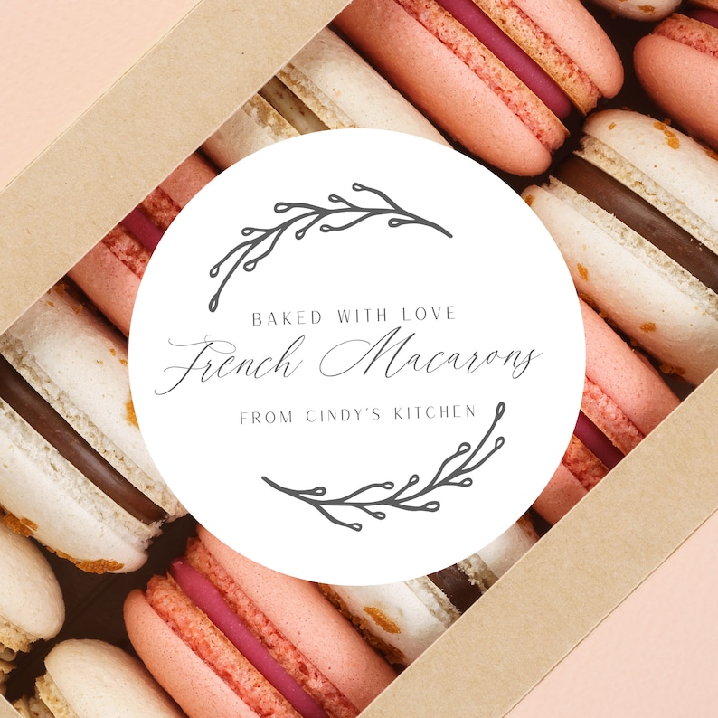 May include: A box of French macarons in various colors, including pink, white, and brown. The macarons are arranged in rows inside a cardboard box. A white circular label with the text "Baked with Love French Macarons From Cindy's Kitchen" is in the center.