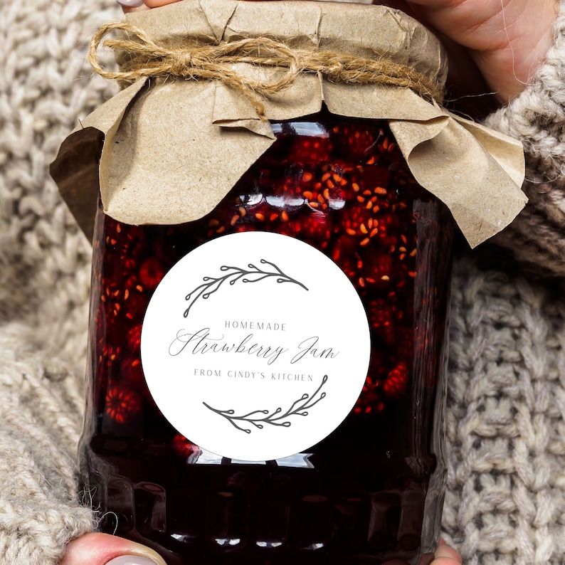 May include: A jar of homemade strawberry jam with a brown paper lid and twine tie. The jar has a white label with the words "Homemade Strawberry Jam From Cindy's Kitchen" in a decorative font, surrounded by a floral design.